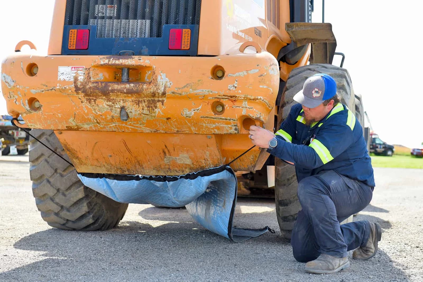 Rear view of the actual Spill Diaper beneath a loader while an operator secures a corner hook
